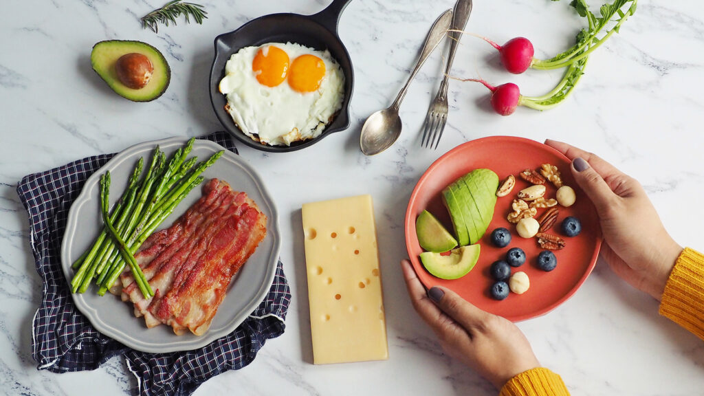 Imagine cu farfurii cu mâncare: avocadă cu nuci și afine, bacon cu sparanghel, ouă, brânză, așezate pe o masă din marmură.