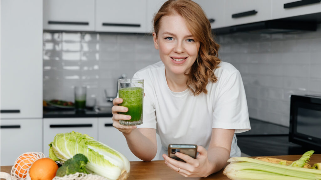 femei zambeste la camera, in bucatarie, bea un smoothie sanatos in timp ce isi calculeaza pe telefon caloriile