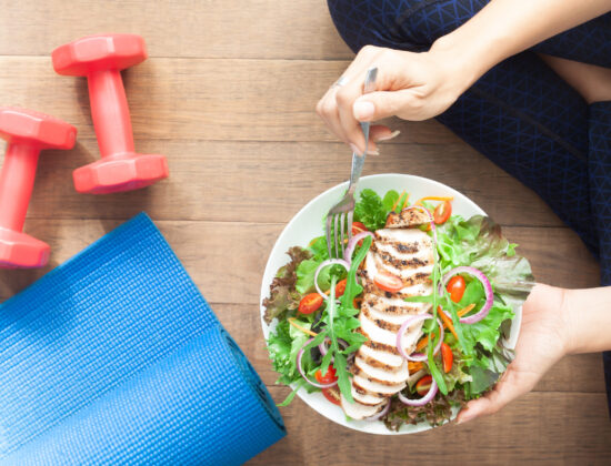 Imagine cu vedere de sus, cu o femeie care mănâncă o salată cu pui. Ea stă pe podea, iar în jurul ei se află echipament sportiv: gantere și covor de yoga