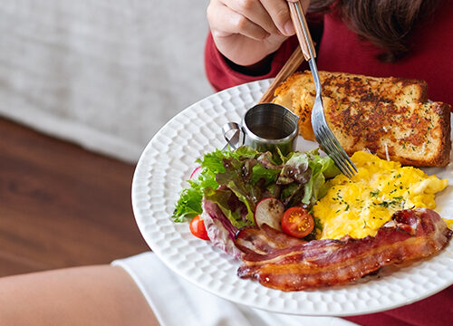 Imagine cu femeie stând pe podea, cu o farfurie având o omletă, o felie de pâine, bacon și salată.