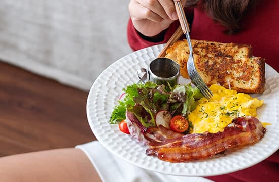 Imagine cu femeie stând pe podea, cu o farfurie având o omletă, o felie de pâine, bacon și salată.