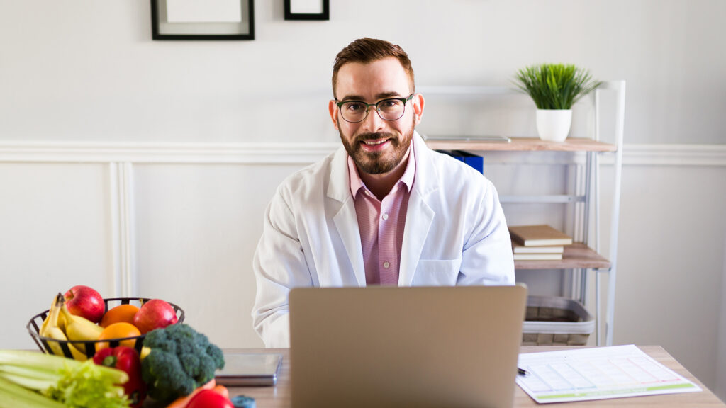 Nutritionist zambeste la camera, in cabinet, in fata laptopului cu alimente santaose pe masa