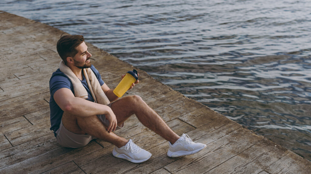 Barbat sportiv, bea apa la rasarit dupa antrenament, la marginea lacului