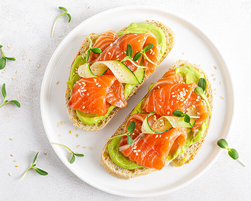 Doua felii de toast cu somon si avocado pe farfurie alba pe masa