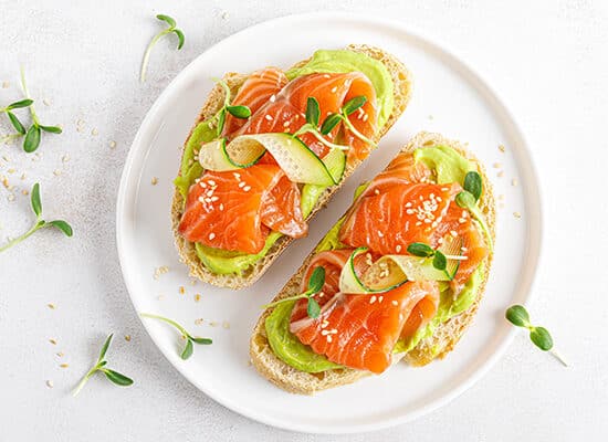 Doua felii de toast cu somon si avocado pe farfurie alba pe masa