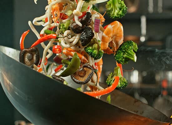 Imagine cu un wok cu creveți și legume ținut în mână de un bucătar.
