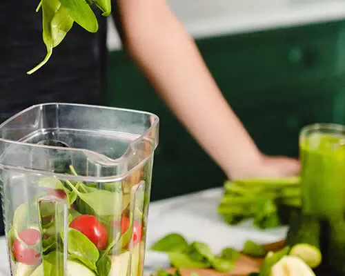 Fotografie cu o femeie stând lângă un blat de bucătărie pe care sunt așezate fructe și legume proaspete, pregătind un smoothie.