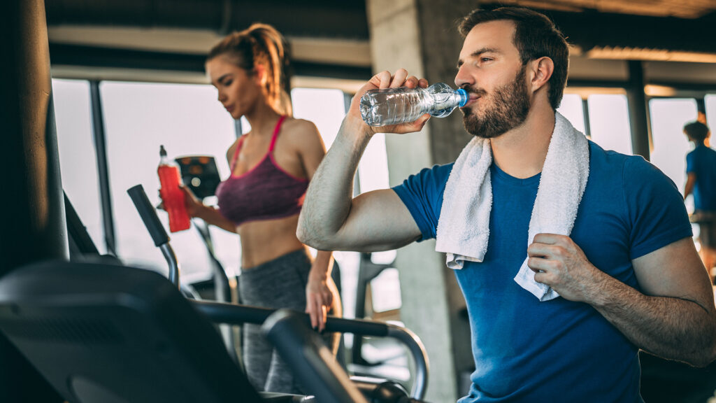 Barbat si femeie in sala de fitness, alearga pe banda in timp ce beau apa