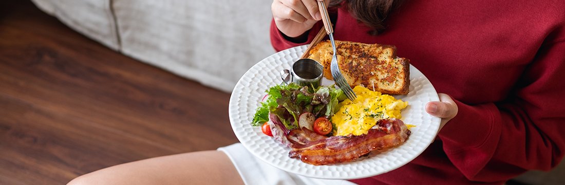 Imagine cu femeie stând pe podea, cu o farfurie având o omletă, o felie de pâine, bacon și salată.