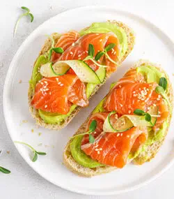 Doua felii de toast cu somon si avocado pe farfurie alba pe masa