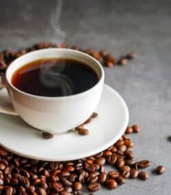 cafea fierbinte in ceasca alba, iese abur din ea, boabe de cafea in jur