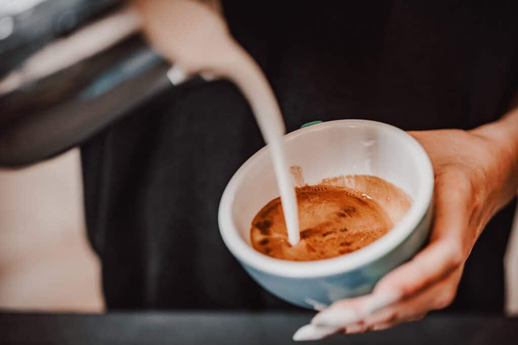 Imagine de prim plan ce surprinde mâinile unui barista care toarnă lapte într-o ceașcă albastră de cafea 