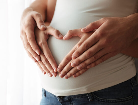 Femeie însărcinată cu mânuțele pe burtică, îmbrățișată de partenerul ei