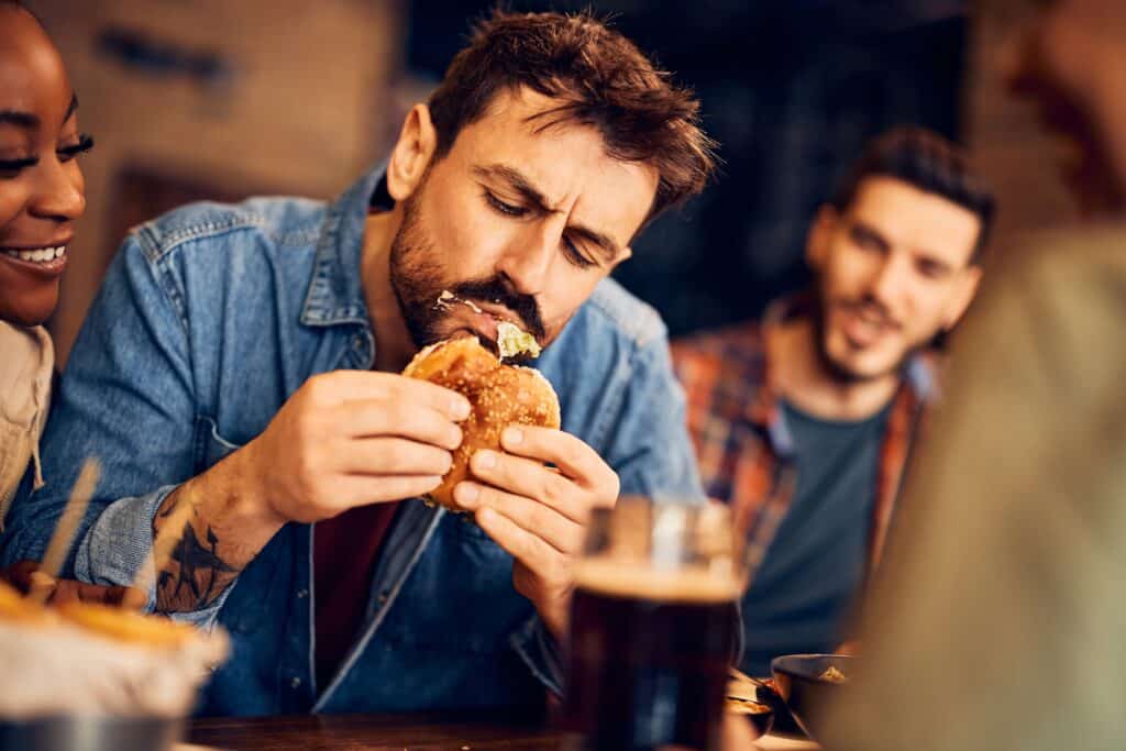 Bărbat înfometat care mănâncă burger în timp ce se adună cu prietenii într-un pub.