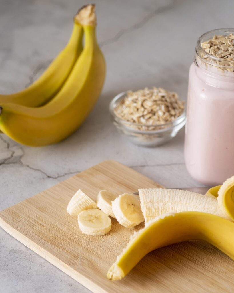 Imagine cu ce sa mananci la mahmureala. Se poate vedea o banană tăiată pe un tocător de lemn. În spate sunt alte două banane întregi și un bol cu iaurt de fructe cu ovăz.