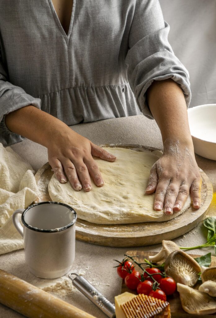 Imagine din față cu mâinile unei femei care întinde aluatul de pizza de casă pe un tocător rotund de lemn. În jur sunt ingrediente folosite pentru o pizza de casă, cum ar fi făina, pentru aluat, dar și roșii, busuioc, ciuperci, cașcaval