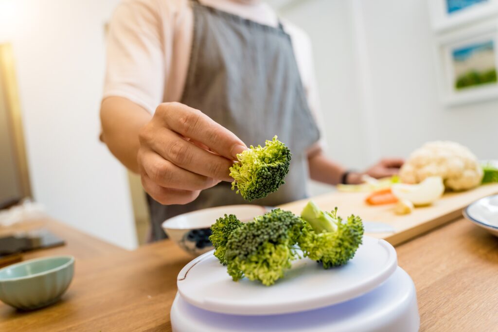 Persoană care prepară legume. Imagine de prim plan cu mâna persoanei care gătește. Aceasta cântărește buchețele de broccoli. În fundal se observă un tocător cu alte legume: morvoc, ceapă, conopidă.