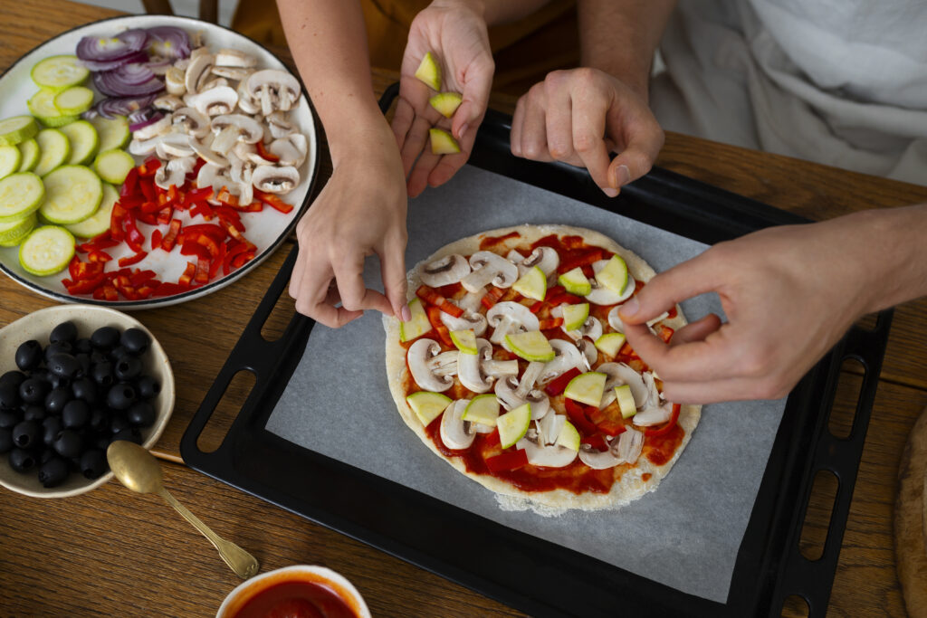 Imagine de aproape cu mâinile a două persoane care pun ingrediente pe un blat de pizza uns cu sos de roșii: ciuperci, dovlecel, ardei gras, ceapă roșie, măsline