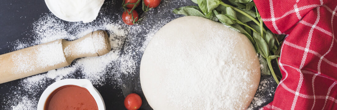 Imagine cu vedere de sus ce arata un aluat de pizza pufos înconjurat de ingrediente precum făină, sos de roșii, roșii cherry, brânză pe un fundal negru. Blatul e presărat cu făină, iar lângă aluat este un prosop roșu, ce arată că aluatul de pizza trebuie lăsat la dospit