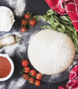 Imagine cu vedere de sus ce arata un aluat de pizza pufos înconjurat de ingrediente precum făină, sos de roșii, roșii cherry, brânză pe un fundal negru. Blatul e presărat cu făină, iar lângă aluat este un prosop roșu, ce arată că aluatul de pizza trebuie lăsat la dospit