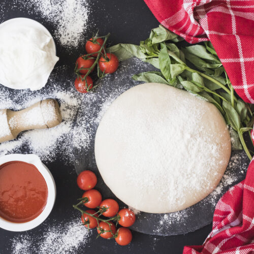 Imagine cu vedere de sus ce arata un aluat de pizza pufos înconjurat de ingrediente precum făină, sos de roșii, roșii cherry, brânză pe un fundal negru. Blatul e presărat cu făină, iar lângă aluat este un prosop roșu, ce arată că aluatul de pizza trebuie lăsat la dospit