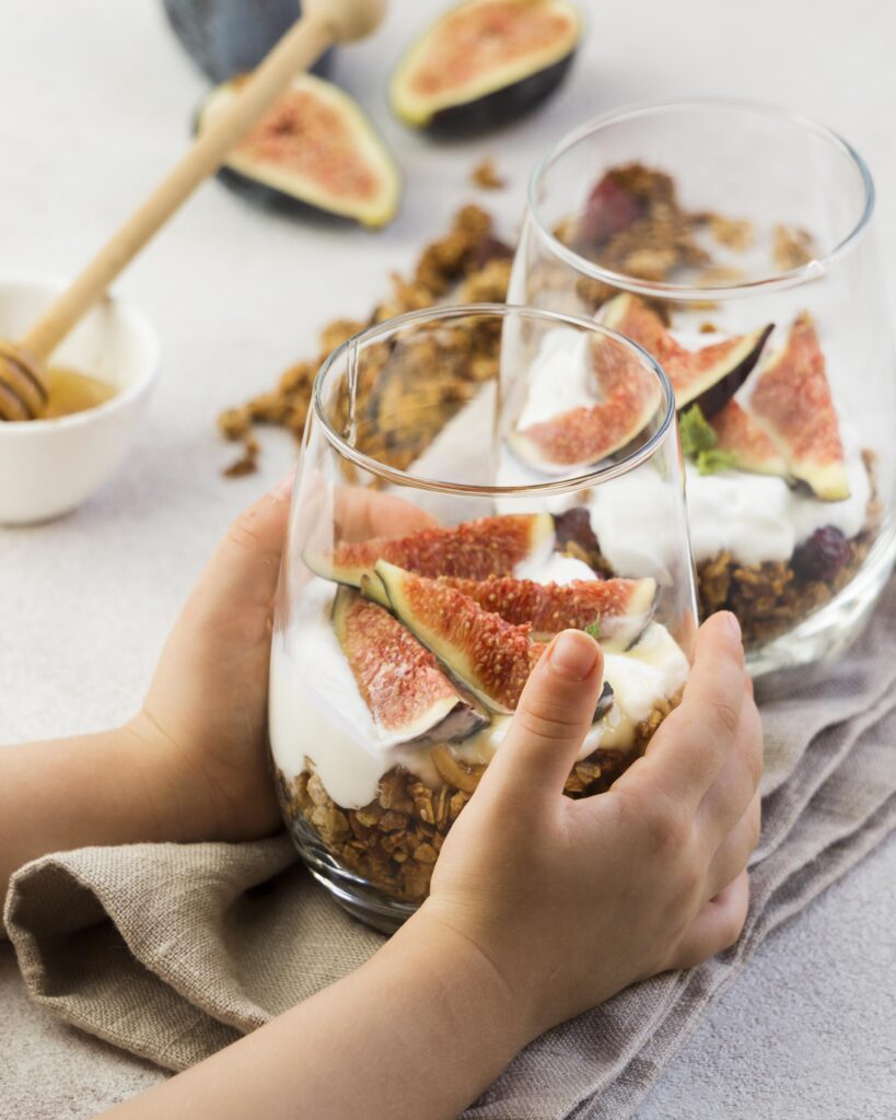 Imagine de aproape cu mâinile unui copilaș careține un bol înalt cu un amestec de granola, iaurt și smochine pe deasupra