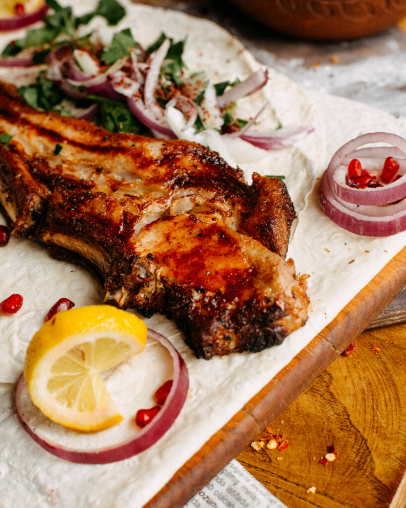 Imagine de aproape cu o bucată de carne de miel pe un tocător de lemn, înconjurată de diferite legume și fructe: salată, ceapă, rodie și o felie de lămâie
