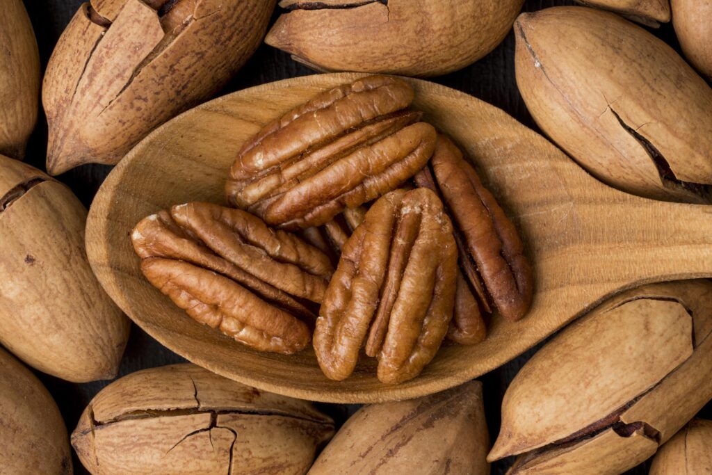 Imagine de aproape cu o lingură de lemn cu miezul unor nuci pecan, pe un fundal de nuci pecan intregi