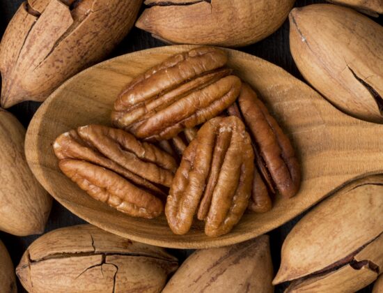 Imagine de aproape cu o lingură de lemn cu miezul unor nuci pecan, pe un fundal de nuci pecan intregi