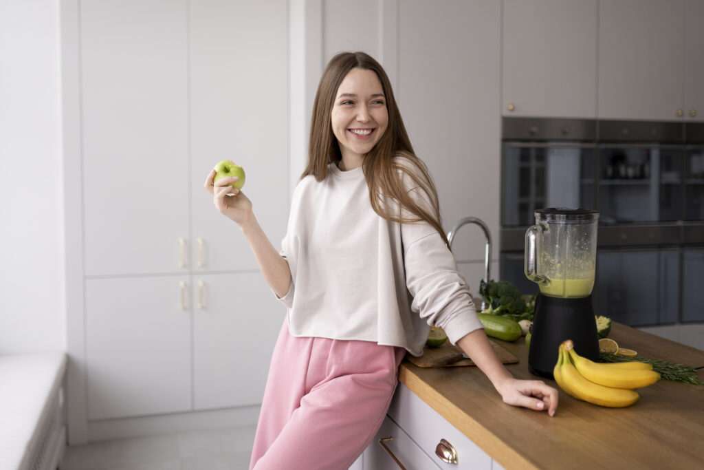 Imagine din fata cu o fata tanara fericita care mananca un mar in timp ce isi pregateste un smoothie de fructe