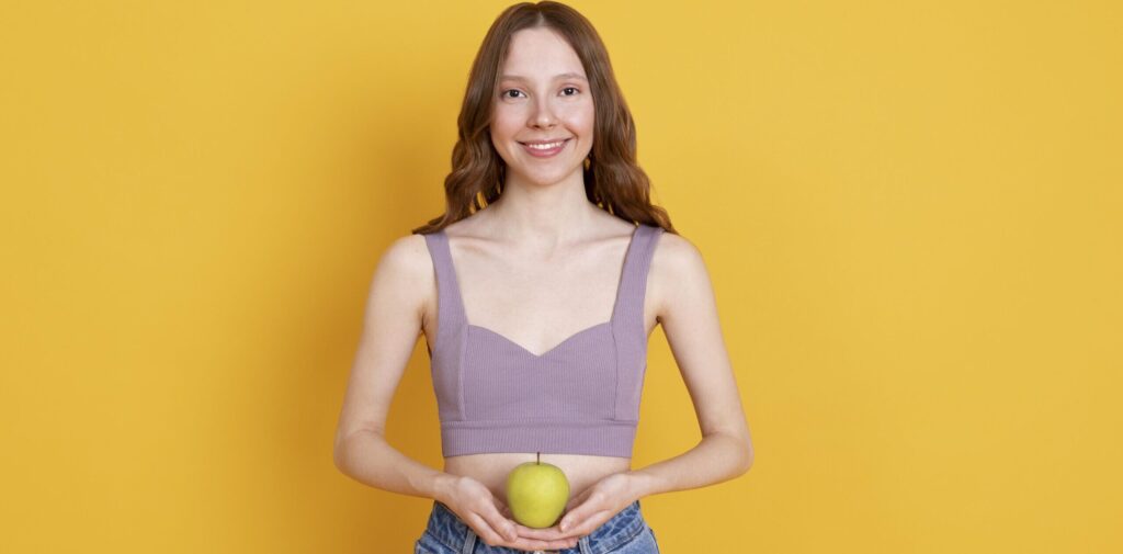 Imagine din față cu o fată tănără ce ține un măr în dreptul abdomenului drept semn pentru un microbiom intestinal sănătos
