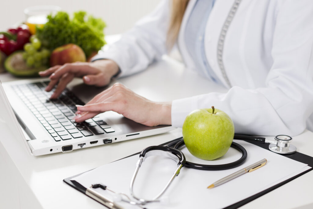 Imagine de aproape cu un medic care tasteaza la laptop. Langa, un stetoscop si un mar. In fundal se vad numeroase fructe si legume