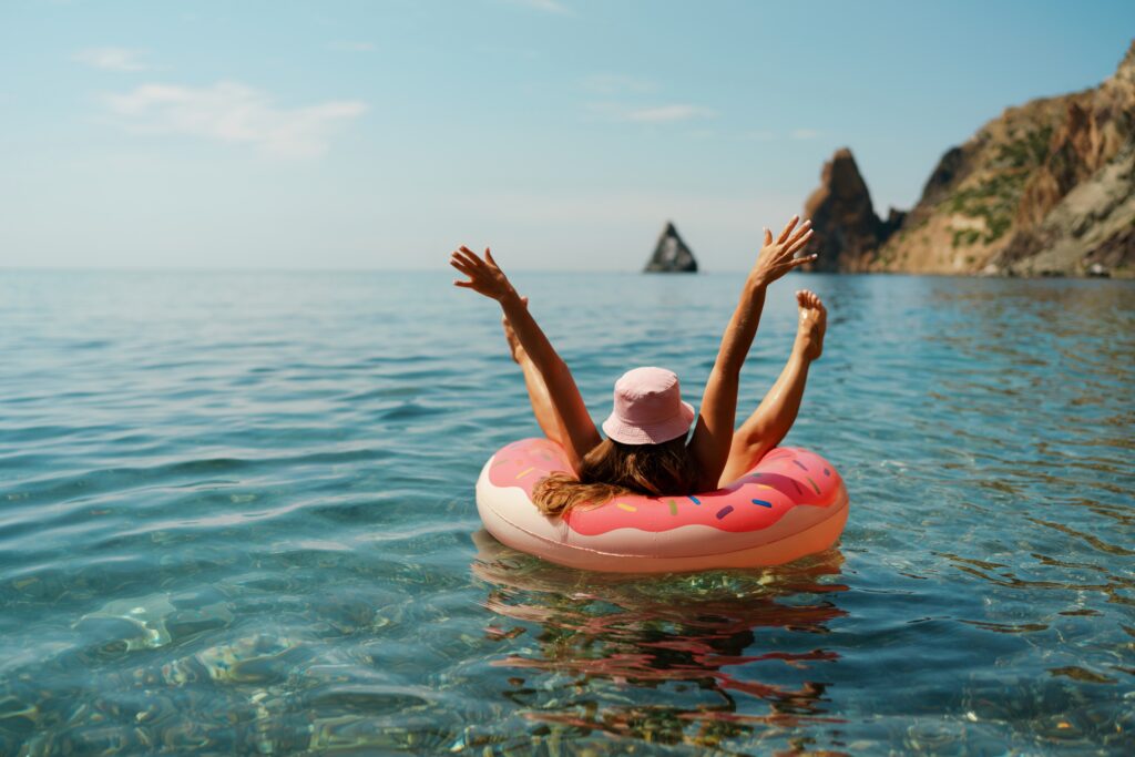 femeie pluteste pe o saltea gonflabila pe mare, in vacanta, cu o palarie pe cap si cu mainile in aer