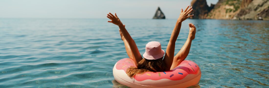 femeie pluteste pe o saltea gonflabila pe mare, in vacanta, cu o palarie pe cap si cu mainile in aer