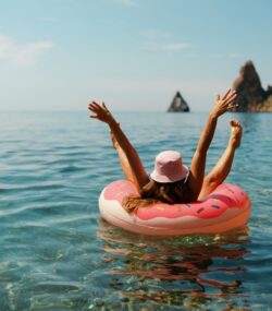 femeie pluteste pe o saltea gonflabila pe mare, in vacanta, cu o palarie pe cap si cu mainile in aer