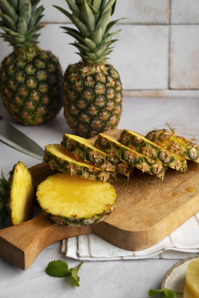 Rondele de ananas pe un tocator de lemn. In spate, 2 ananasi intregi, aliment optim pentru digestie sanatoasa