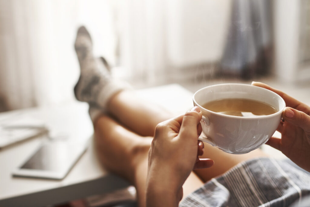 Imagine de aproape cu o persoana care tine in maini o ceasca cu cafea si o bea de dimineata, in pijamale