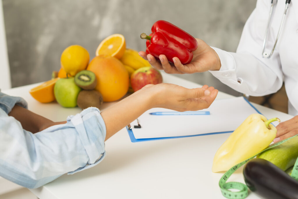 Imagine de aproape cu mana unui medic nutritionist care ii da pacientului un ardei gras. in spate, diferite fructe si legume, pe masa