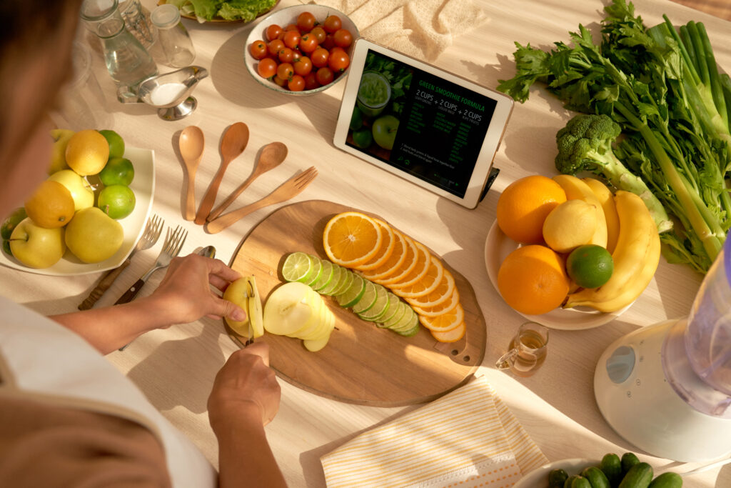 Imagine cu vedere de sus, femeie care taie fructe pe un tocator de lemn, urmareste pe tableta o reteta de smoothie