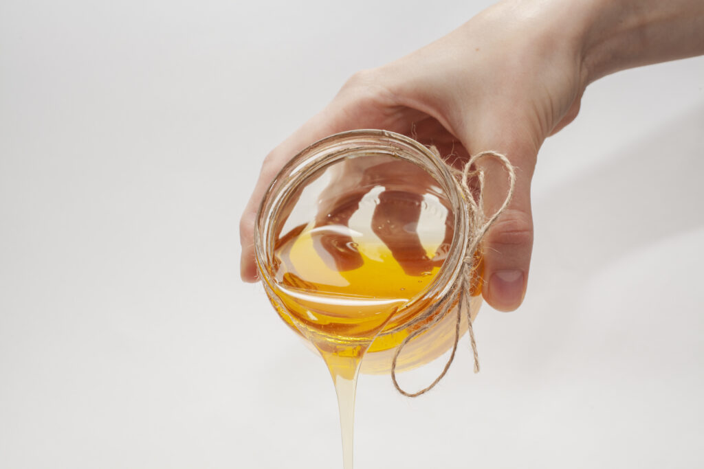 Mâna unei persoane care toarnă sirop de agave dintr-un borcănel transparent, pe un fundal alb