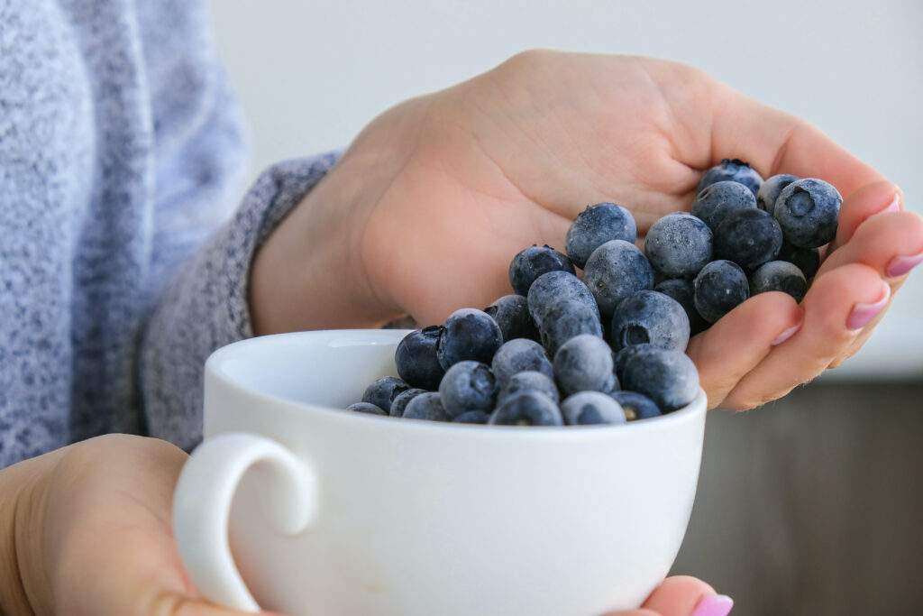 Imagine de aproape cu mainile unei femei. In una tine o canuta alba cu afine, in cealalta tine alte afine, alimente sanatoase cu beneficii antioxidante