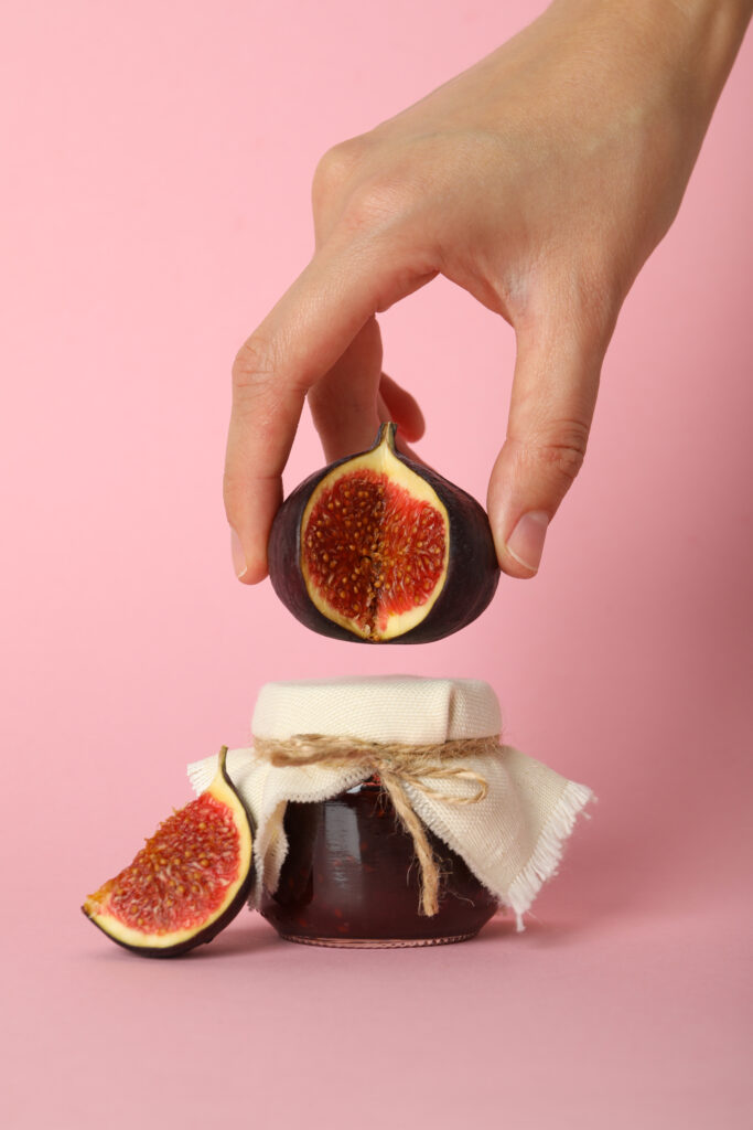 Imagine de aproape. Mana unei persoane care tine o smochina deasupra unui borcanel cu dulceata de smochine. Fundal rozc