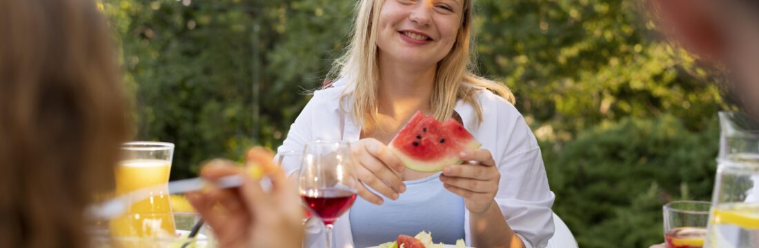 Femeie tanara, blonda, se uita catre camera zambind cu o felie de pepene rosu in maini la un picnic in gradina cu prietenii