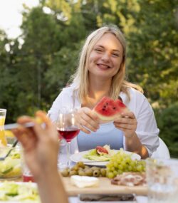 Femeie tanara, blonda, se uita catre camera zambind cu o felie de pepene rosu in maini la un picnic in gradina cu prietenii