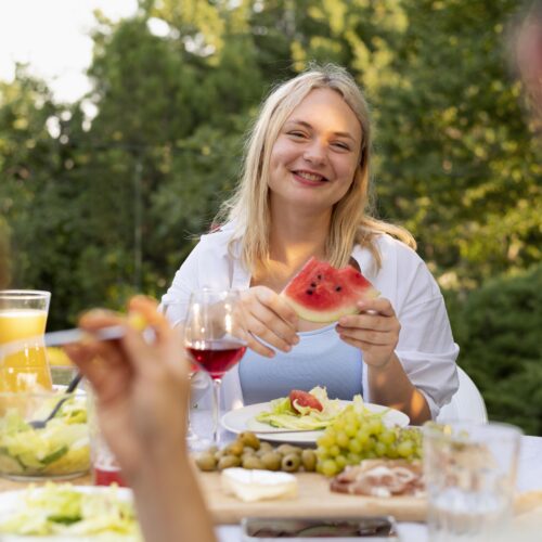 Femeie tanara, blonda, se uita catre camera zambind cu o felie de pepene rosu in maini la un picnic in gradina cu prietenii