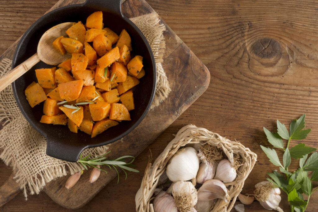 Craticioara cu cartofi dulci copti, pe un tocator de lemn, langa un cosulet cu usturoi. Cadru rustic