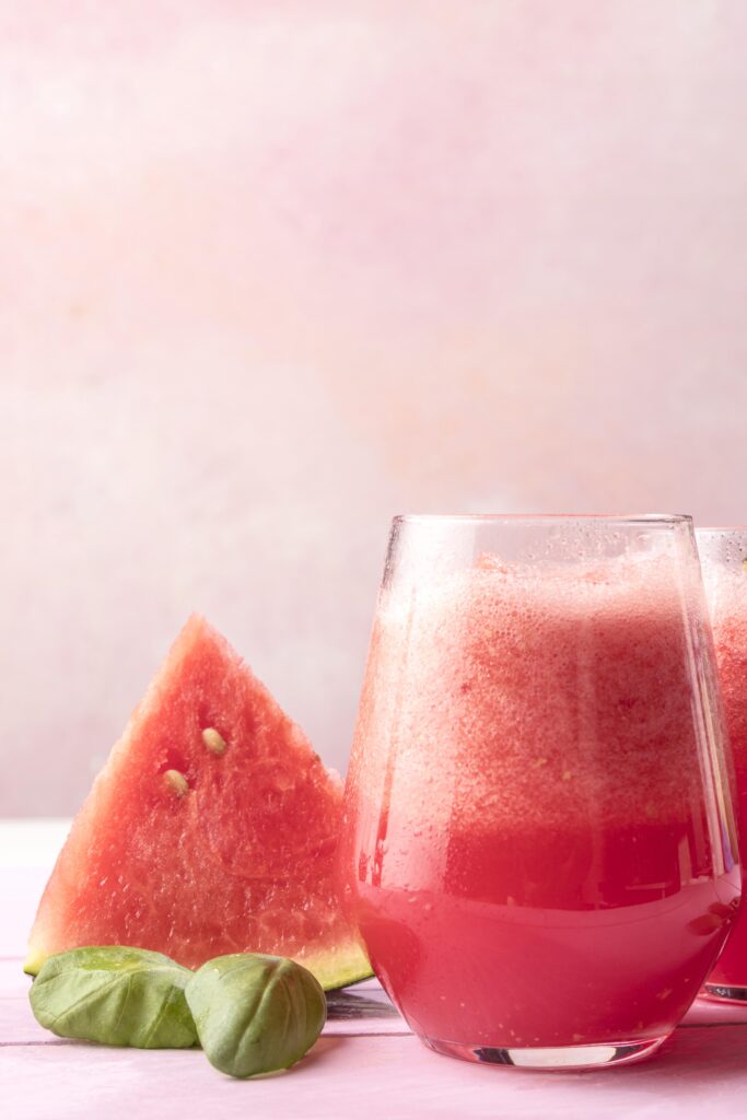 Imagine de aproape cu un pahar de smoothie in dieta detoxifiere cu pepene rosu. Langa, un triunghi de pepene si frunze de menta. Fundal roz