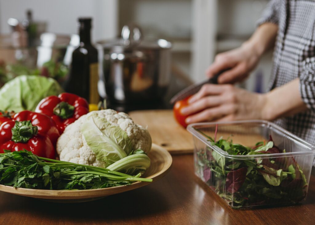 Imagine in fundal cu o persoana care toaca legume pe un tocator de lemn. In fata se observa alte alimente vegetale: salată, ardei gras, rosu, conopidă