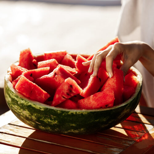 un bol facut din coajă de pepene plin cu bucăți mici de pepene rosu, mana unei femei care ia o bucată de pepene