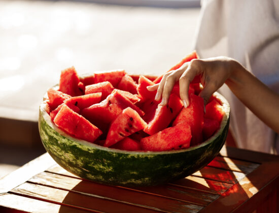 un bol facut din coajă de pepene plin cu bucăți mici de pepene rosu, mana unei femei care ia o bucată de pepene